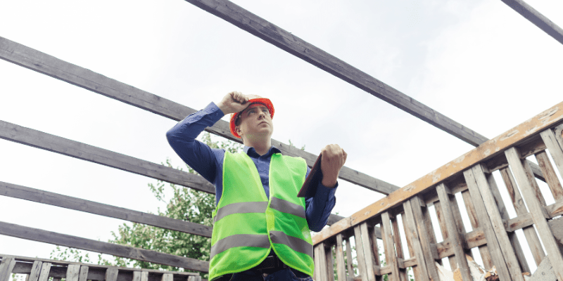 Un professionnel du bâtiment portant un casque et un gilet fluorescent inspecte une structure en bois pendant une levée de réserves.
