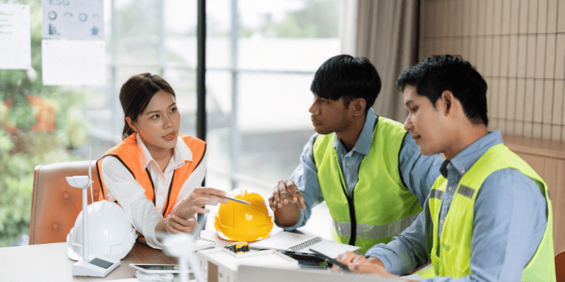 Trois professionnels du BTP en réunion de chantier, discutant autour d'une table avec plans et documents.