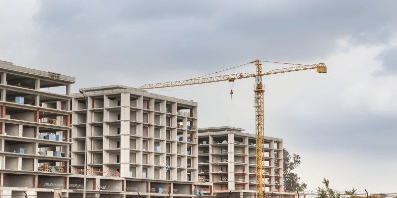 Bâtiment en béton en construction avec une grande grue jaune sur fond de ciel nuageux.