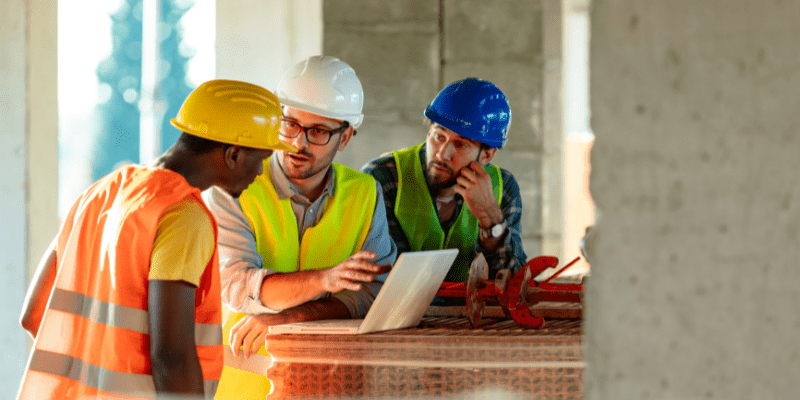 Trois professionnels du BTP (avec casques et gilets de sécurité) vérifiant le PV de levée des réserves et les plans sur un ordinateur portable au milieu du chantier.