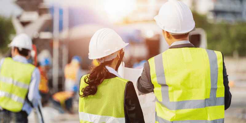 Deux chefs de projet en gilets et casques de sécurité, effectuant le suivi de travaux et vérifiant les plans sur un chantier.