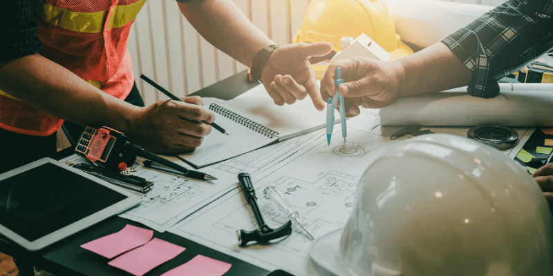 Professionnels du bâtiment discutant autour de plans techniques avec un compas, des carnets et une tablette numérique.