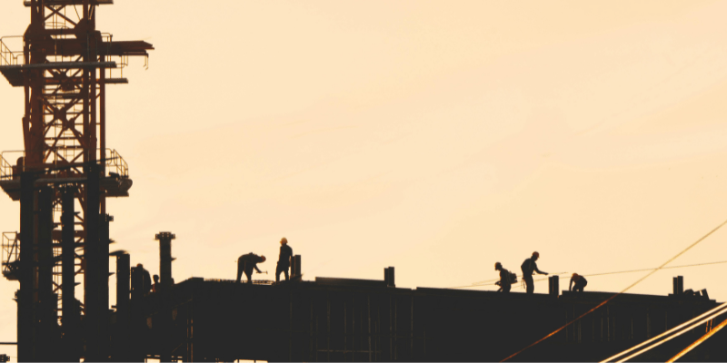 Silhouettes d'ouvriers travaillant sur le sommet d'une structure en construction avec une grue, sur un fond de ciel orangé