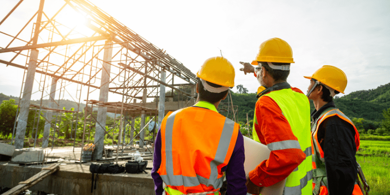 Trois professionnels du bâtiment avec casques et gilets de sécurité inspectant la structure métallique d'un chantier.