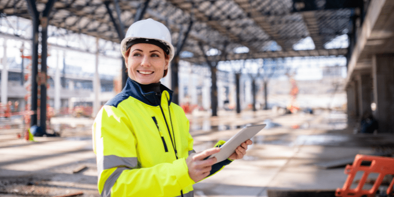 Une femme professionnelle du bâtiment, portant un casque blanc et un gilet haute visibilité, tenant une tablette numérique sur un chantier de construction.