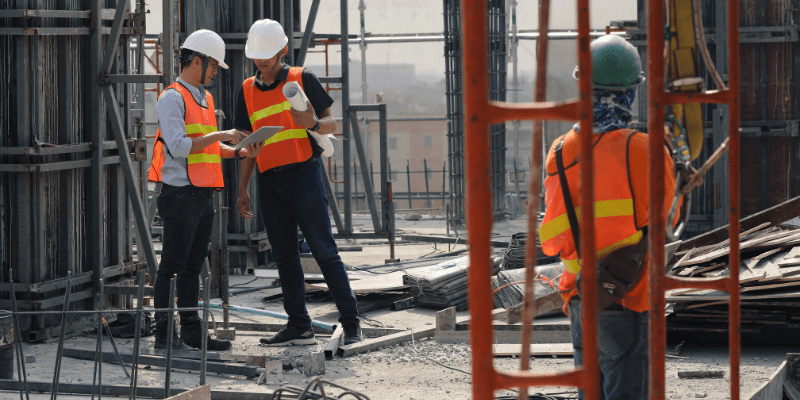 Deux professionnels du bâtiment en gilet de sécurité consultant une tablette numérique sur un chantier de construction.