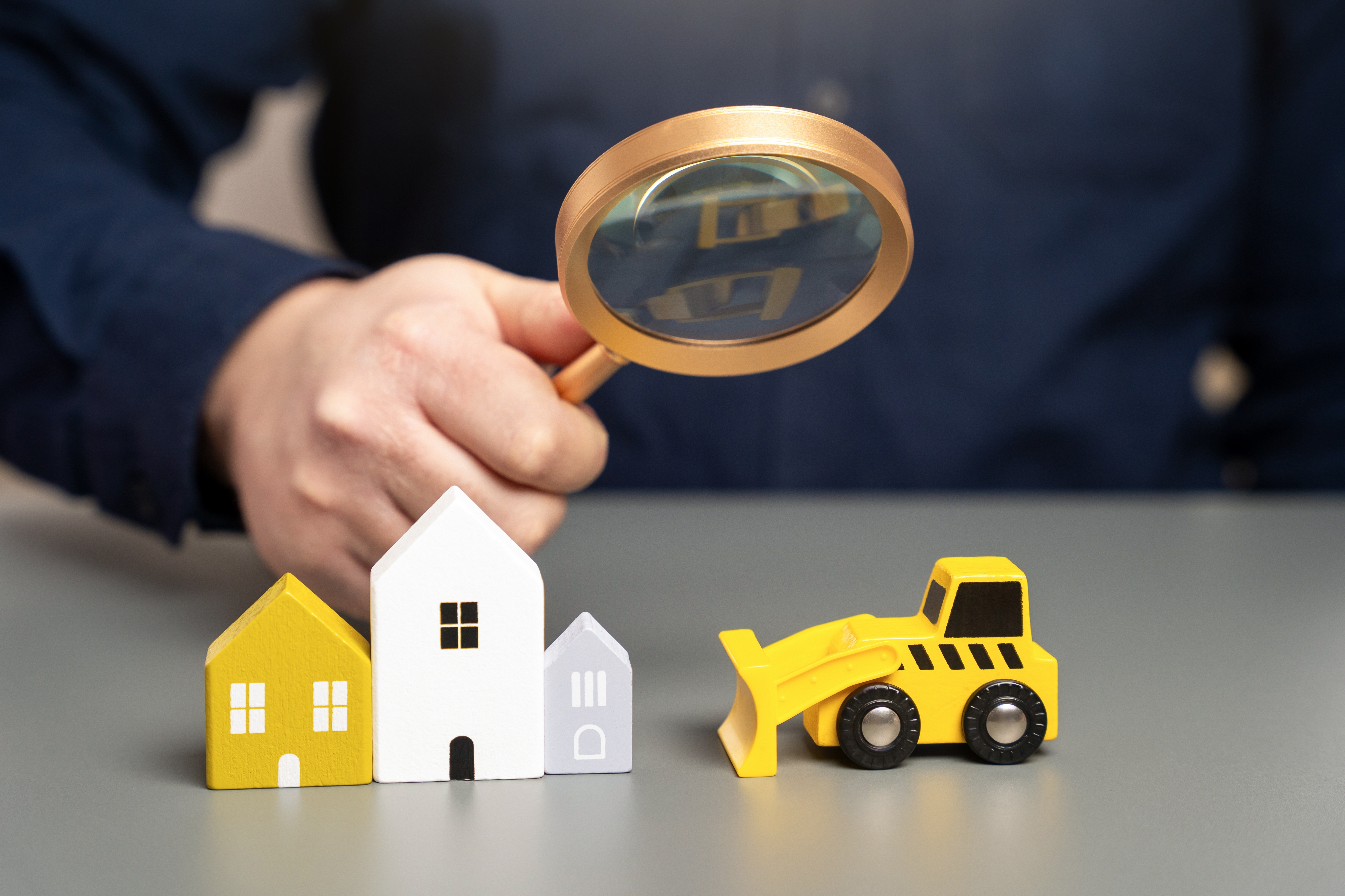 Une main tenant une loupe au-dessus de petites maisons en bois et d'un bulldozer jouet, illustrant l'analyse et la transformation du secteur de la construction.