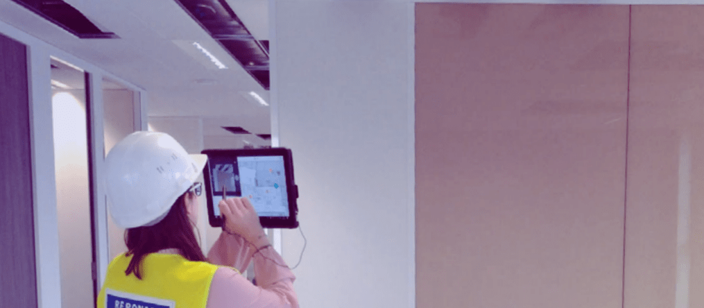 Femme ingénieure portant un casque de chantier et un gilet de sécurité utilisant une tablette pour le suivi de travaux en intérieur.