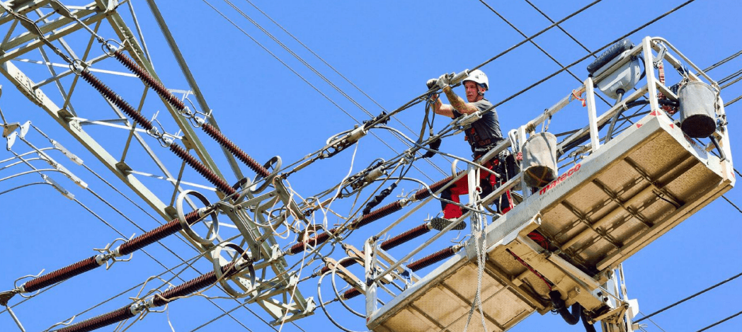 Un technicien EQOS travaillant en hauteur sur une ligne à haute tension depuis une nacelle élévatrice sous un ciel bleu.
