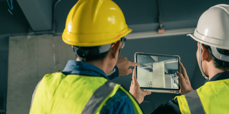 Deux professionnels du BTP en gilets de sécurité et casques de chantier consultent une maquette numérique sur tablette pour inspecter un pilier en béton sur site.