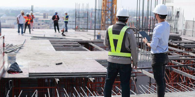 Deux professionnels en équipement de sécurité, dont un avec une tablette, supervisent un chantier en construction avec des structures métalliques en arrière-plan.