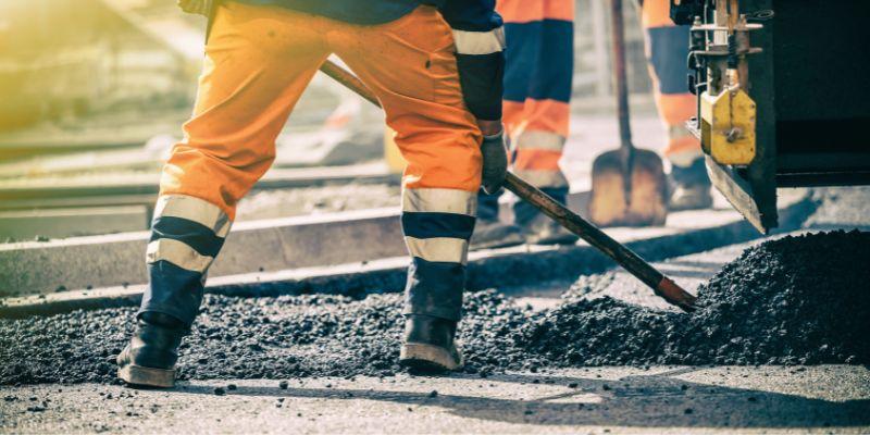 uvriers en tenue de sécurité orange étalant du bitume lors de travaux de voirie sur un chantier routier.