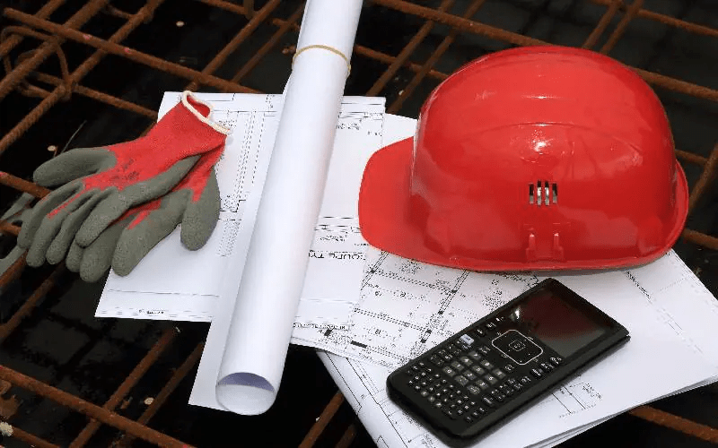 Casque de chantier rouge, gants de protection et plans d'architecte sur un treillis métallique.